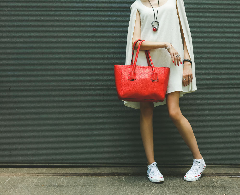 Fashionable beautiful big red handbag on the arm of the girl in a fashionable white dress and sneakers posing near the wall on a warm summer night. Warm color.